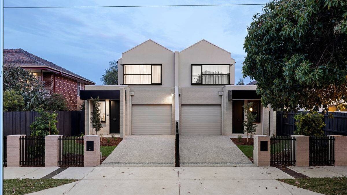 Photograph at dusk of a completed pair of two-storey townhouses with gabled roofs, rendered walls and pale brick, a garage to each.