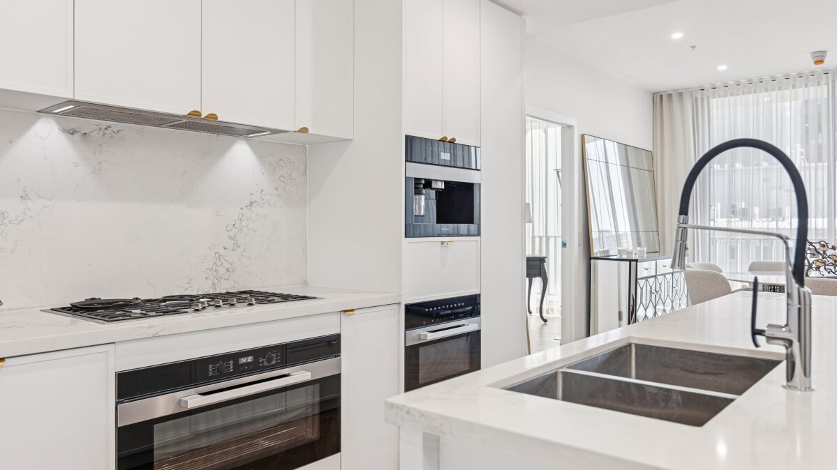 Interior render of a Cullinan apartment kitchen in white and stone, with a gas cooktop, integrated ovens and a window beyond.